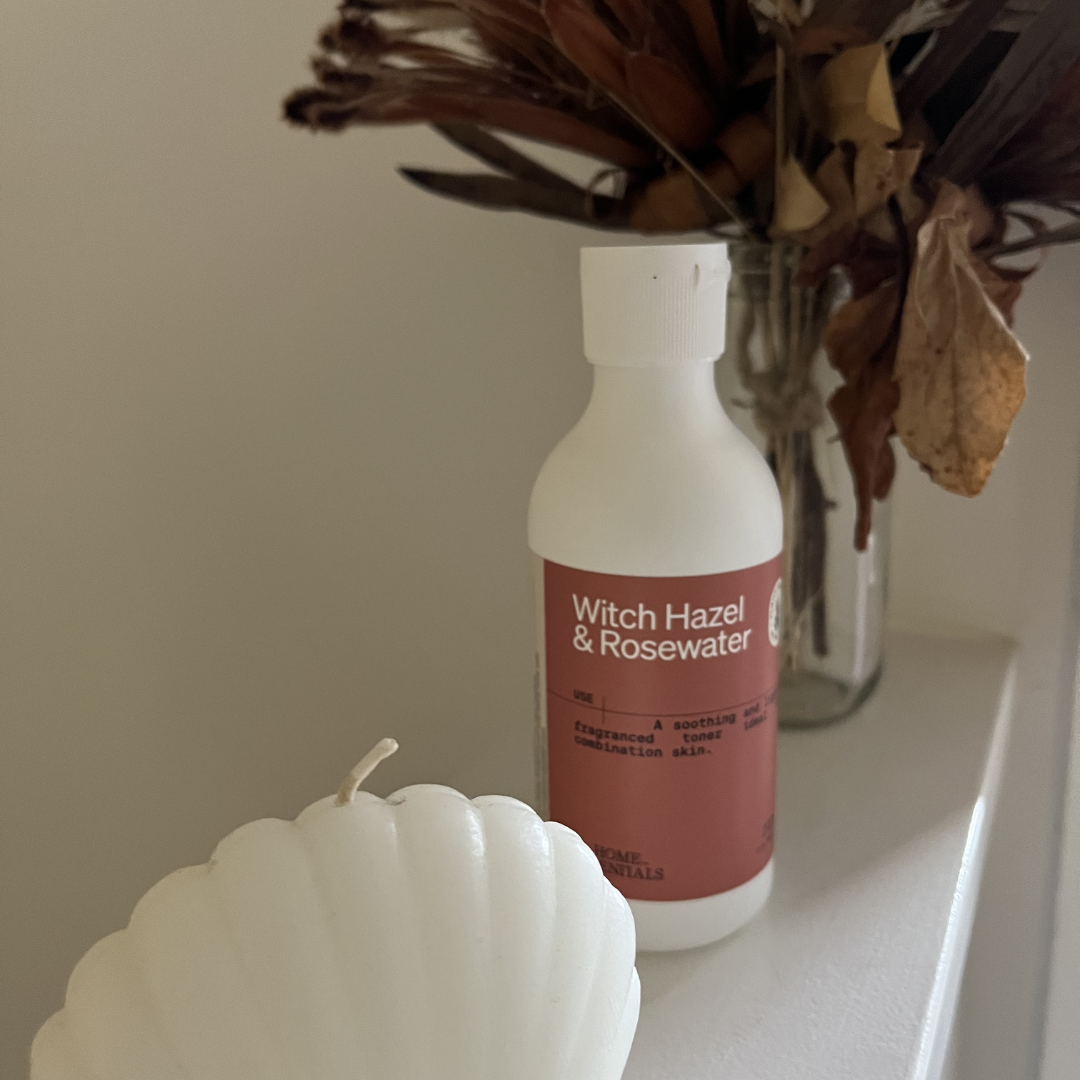 Bottle of Witch Hazel &amp; Rosewater next to a white candle and dried flower bouquet on a shelf.
